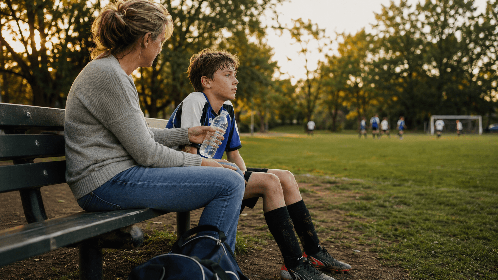 A mother sits close beside her son on a bench at the edge of a soccer field in evening light, the boy looking out at the game instead of at her