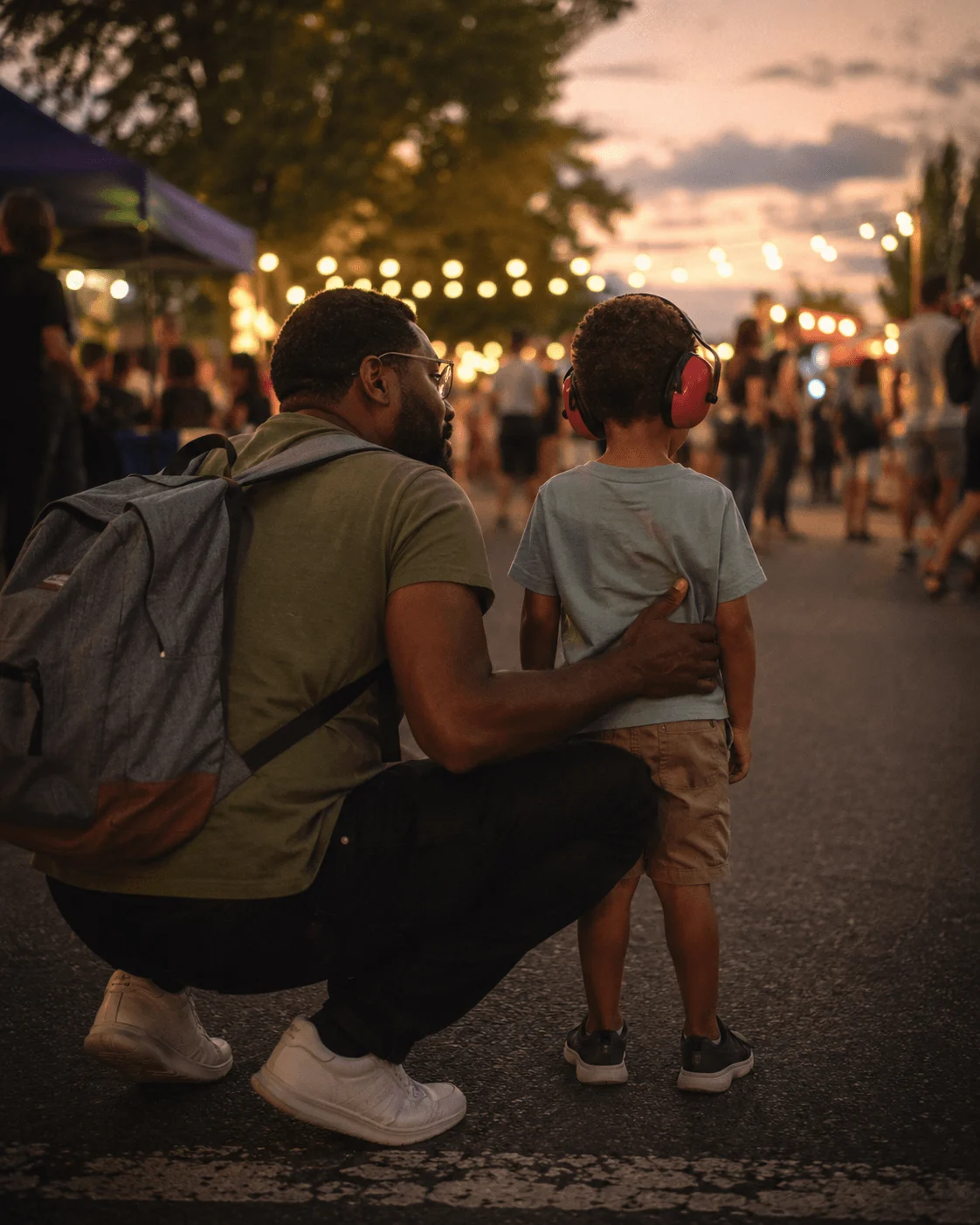 Father holding his son who wears red ear defenders at a crowded outdoor evening event strung with lights