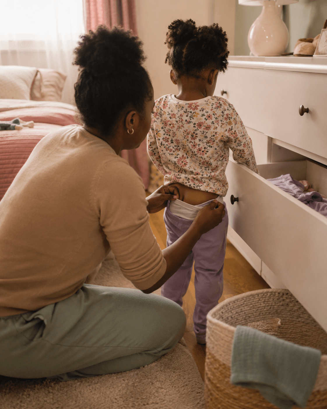 A parent kneels beside a toddler helping guide a leg into clean underwear from an open dresser drawer, both unhurried and matter-of-fact