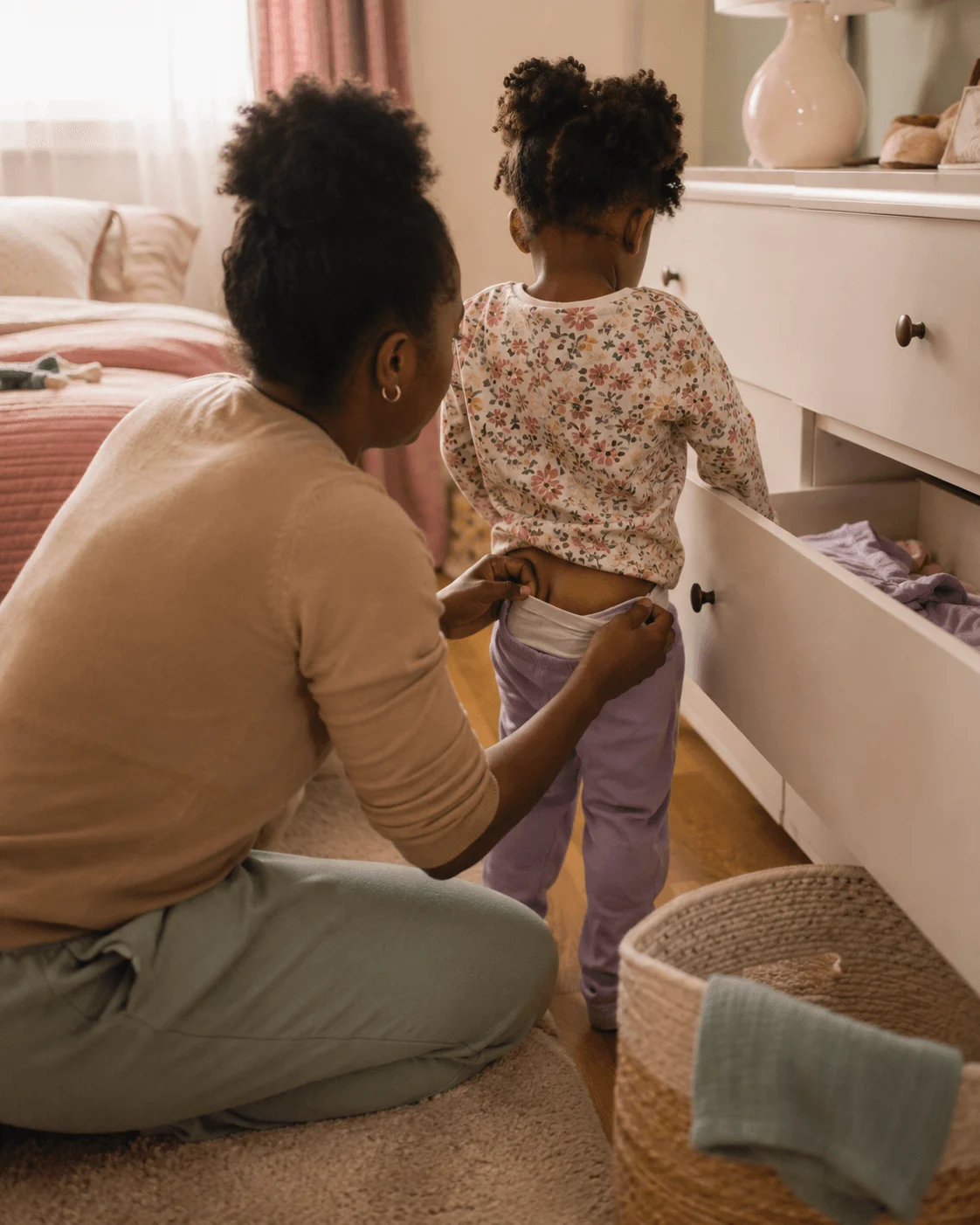 A parent kneels beside a toddler helping guide a leg into clean underwear from an open dresser drawer, both unhurried and matter-of-fact