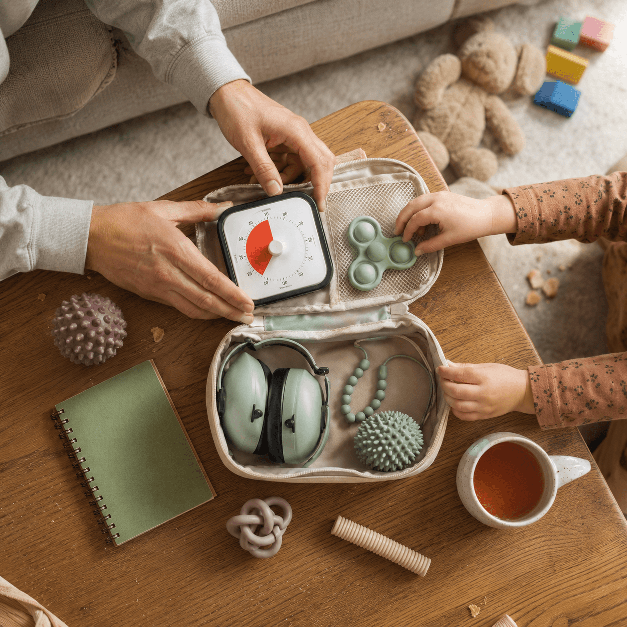 Overhead flat-lay of a sensory kit with a visual timer, ear defenders, chew necklace, popper and textured balls beside a cup of tea