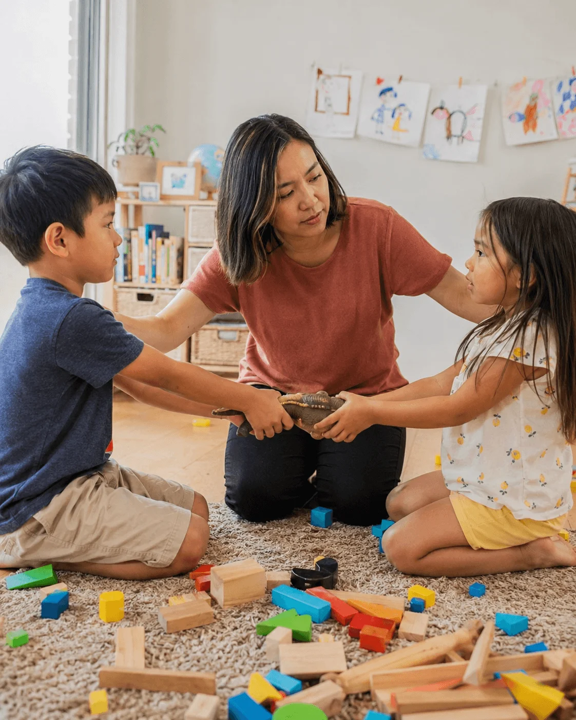 A mother kneels between her son and daughter as they both pull on the same toy dinosaur among scattered wooden blocks