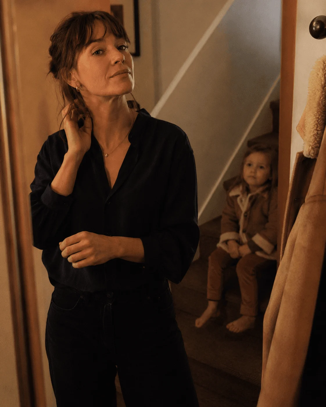 A single mother checks the hallway mirror as she gets ready to go out, her child watching from the stairs