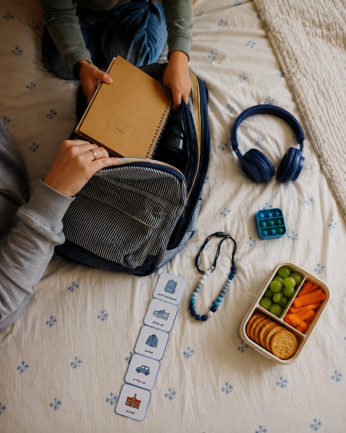 Child packing a backpack with headphones and a visual schedule on the bed