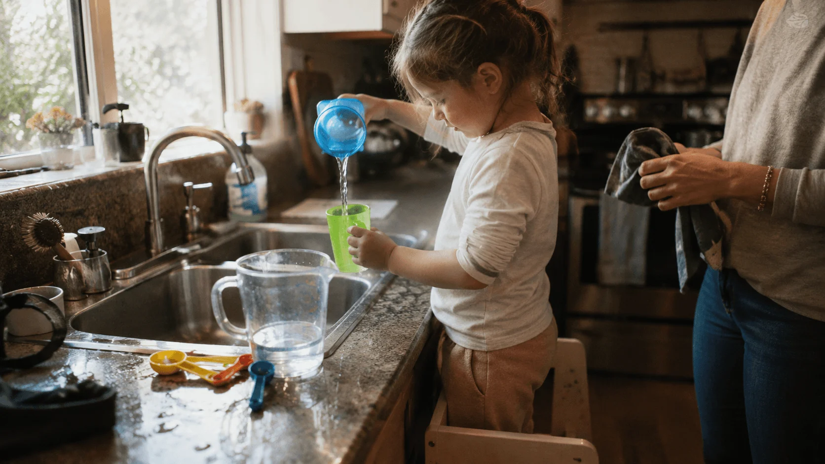 A toddler stands safely in a wooden learning tower at the kitchen sink, pouring water between cups while helping at counter height
