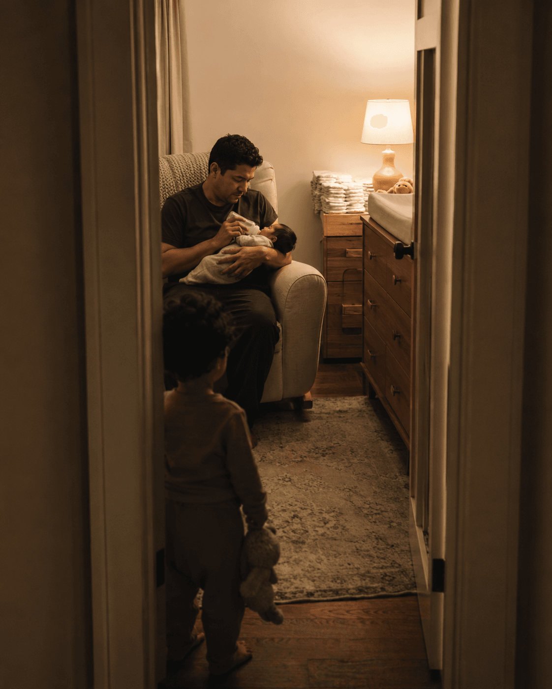 A parent feeds a newborn baby in a nursery chair while an older toddler sibling stands quietly in the doorway watching, holding a stuffed toy