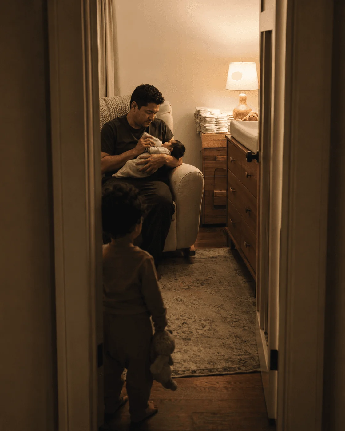 A parent feeds a newborn baby in a nursery chair while an older toddler sibling stands quietly in the doorway watching, holding a stuffed toy