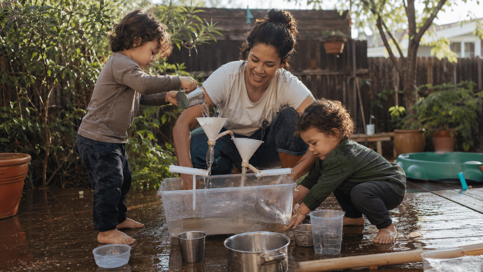 Two toddlers play with a water funnel and cups in a simple backyard setup, absorbed and happy