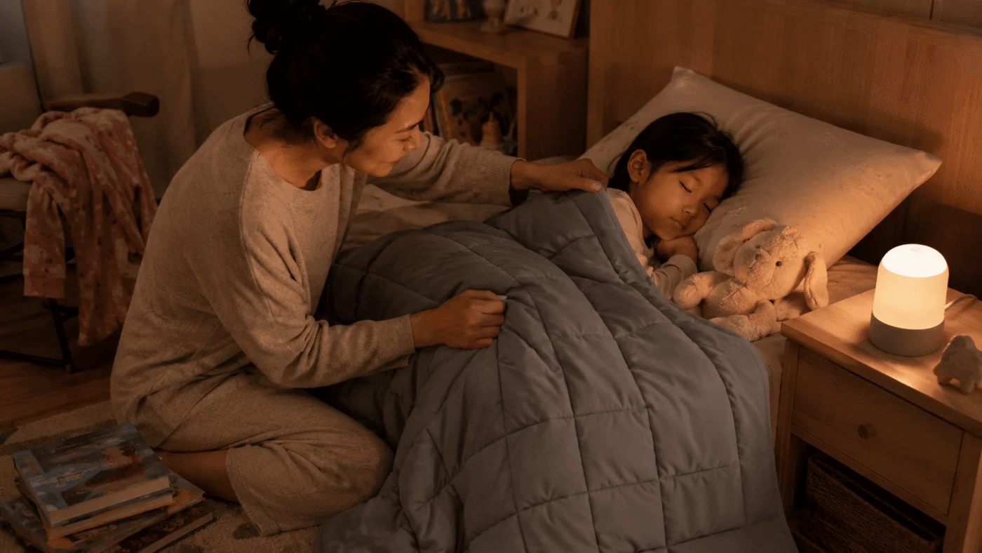 Mother tucks her daughter in under a weighted blanket at bedtime with a bunny and a warm nightlight