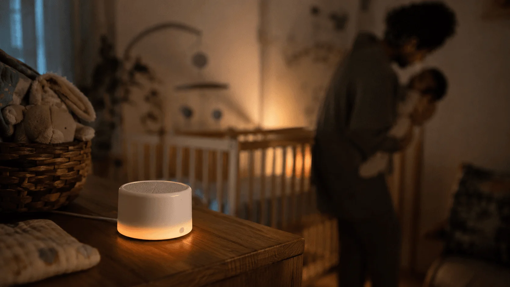 Yogasleep Hushh white noise sound machine glowing amber on a wooden nursery dresser at night, crib and parent softly out of focus behind