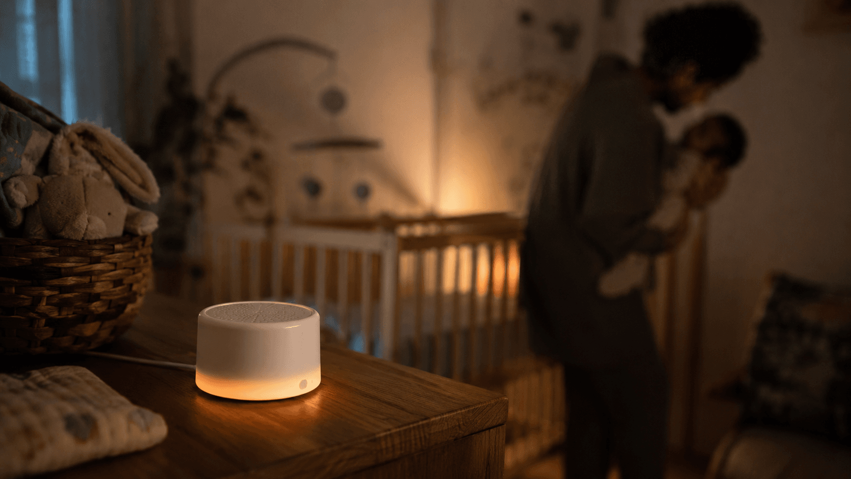 Yogasleep Hushh white noise sound machine glowing amber on a wooden nursery dresser at night, crib and parent softly out of focus behind