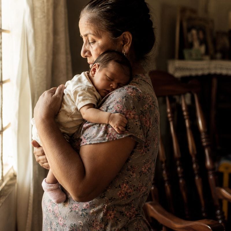 A grandmother holds a newborn by a bright window