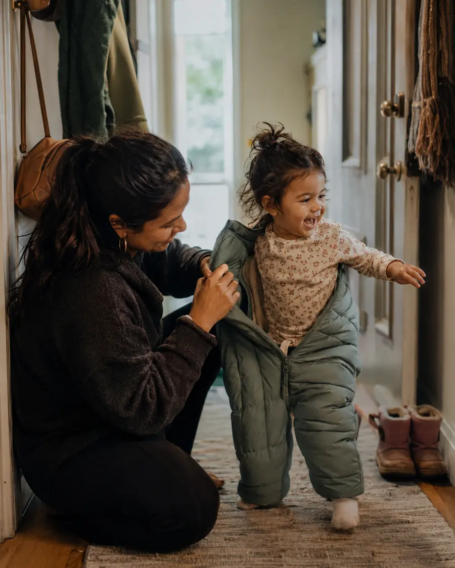 A child struggles into a snowsuit in a hallway while a parent waits