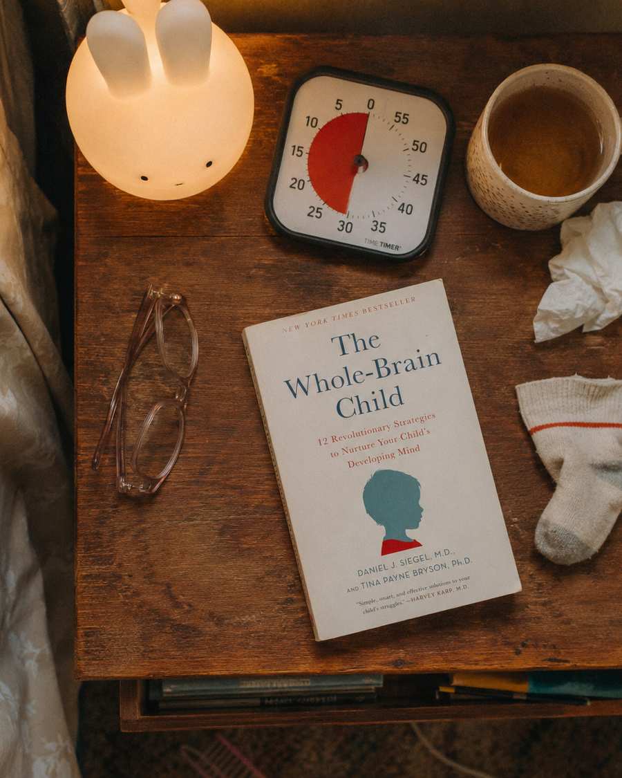 A nightstand with a parenting book, a timer and a small lamp beside a bed