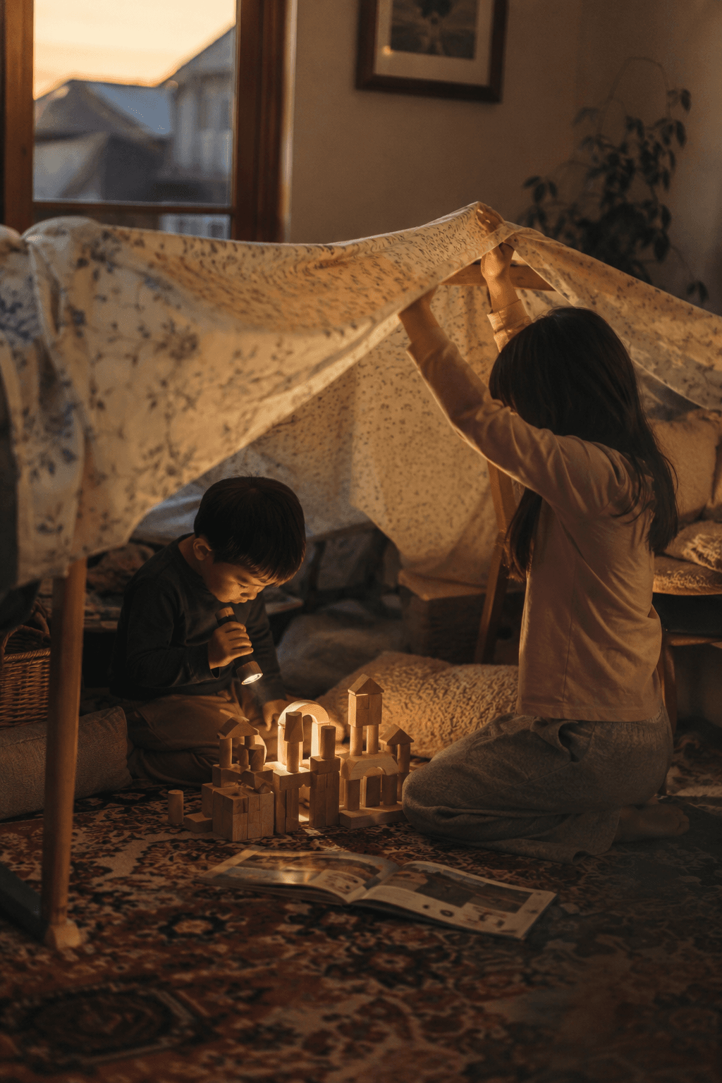 Two kids play in a blanket fort at dusk with a flashlight and a few wooden blocks