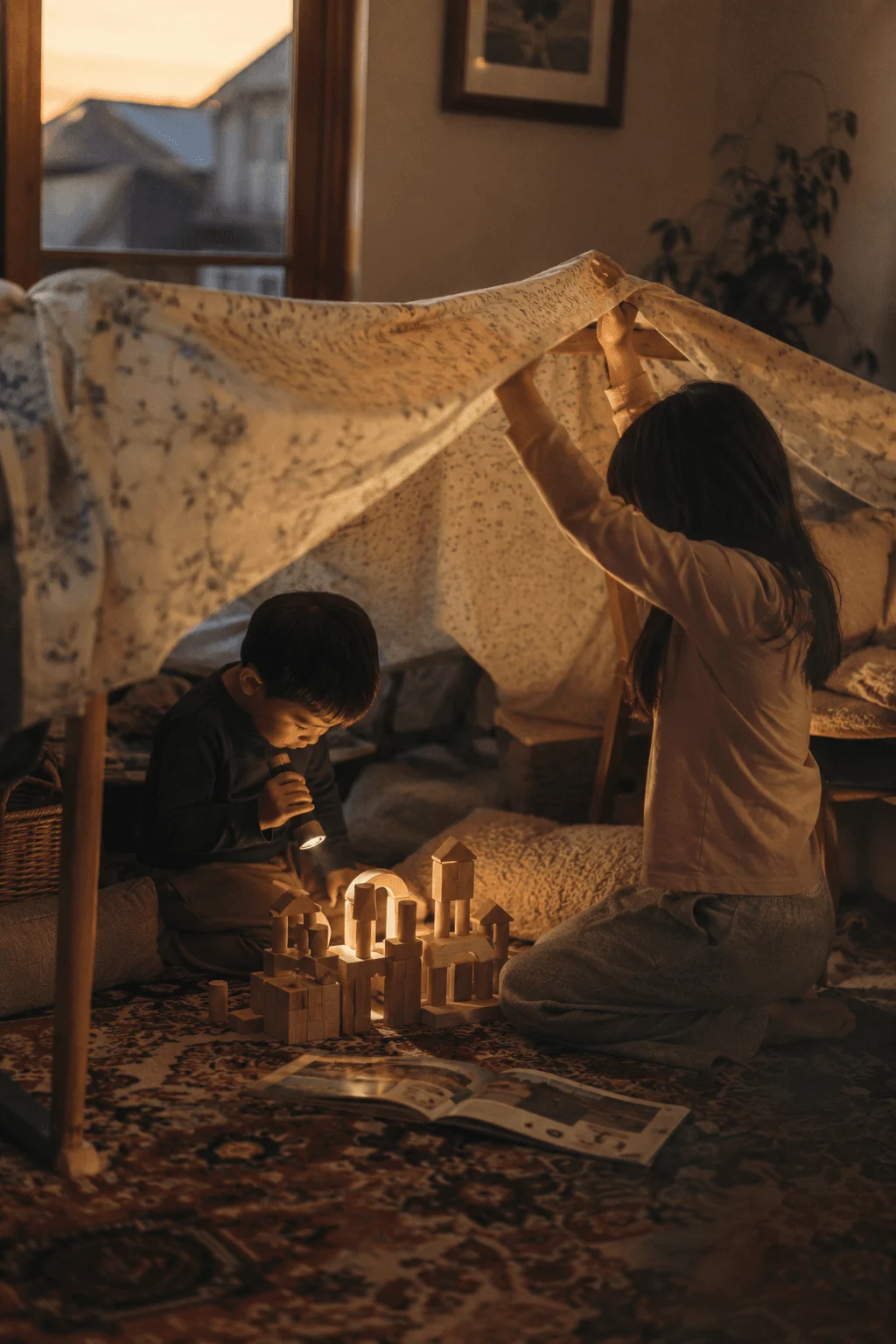 Two kids play in a blanket fort at dusk with a flashlight and a few wooden blocks