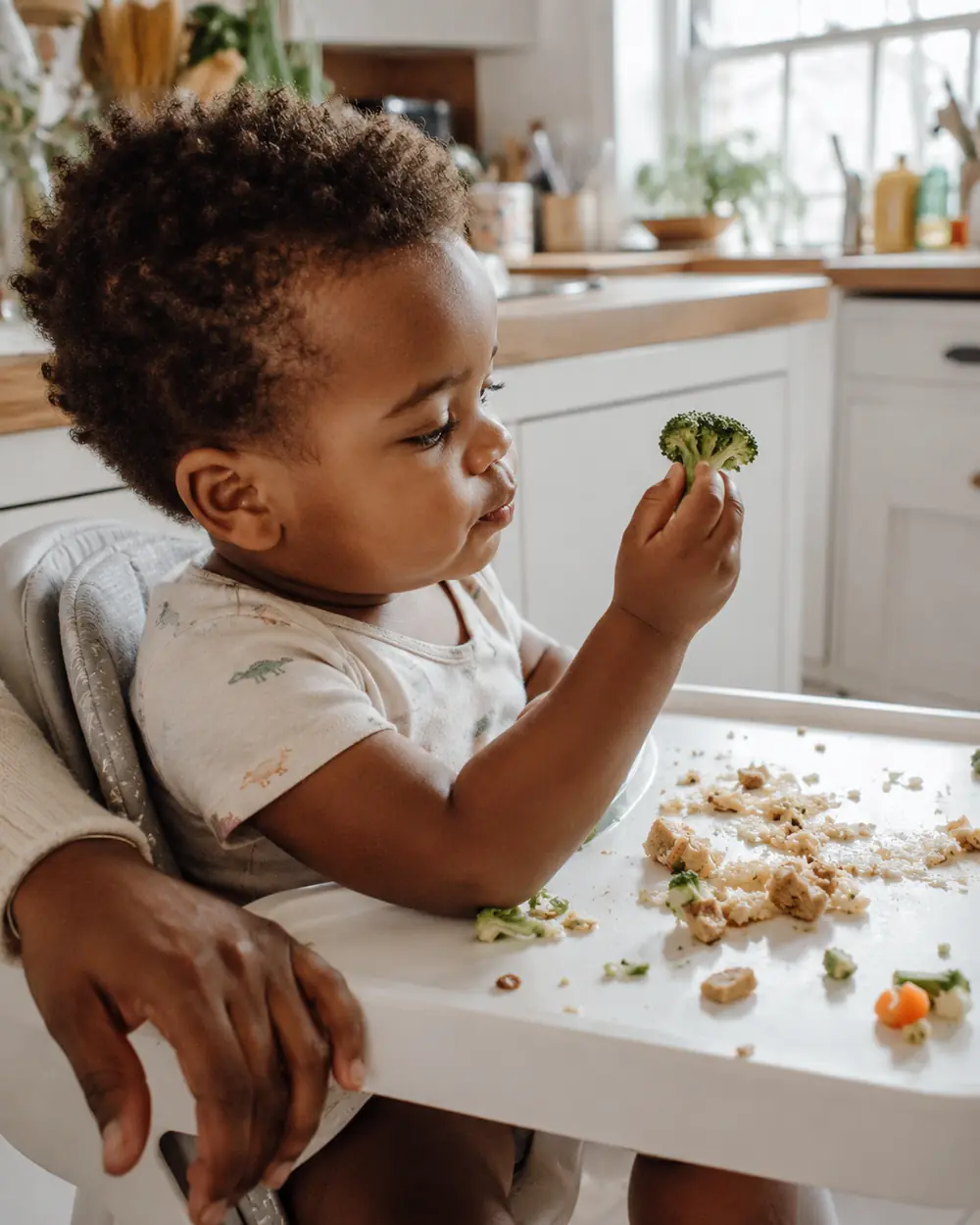 A toddler in a high chair pushes broccoli around the tray instead of eating at dinner time