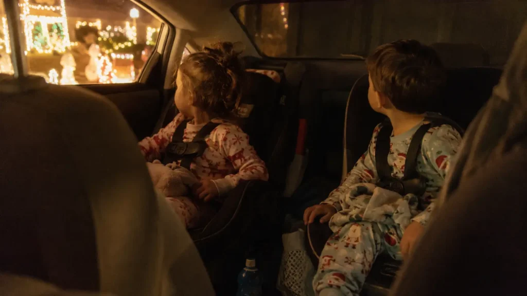 A car interior at night with children in pyjamas looking out at street lights and decorations