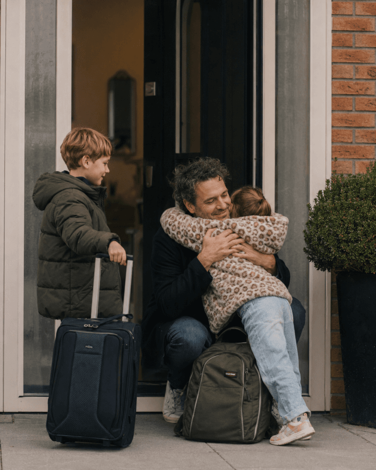 A father kneels in a doorway to hug his daughter while her brother waits beside them with a wheeled suitcase