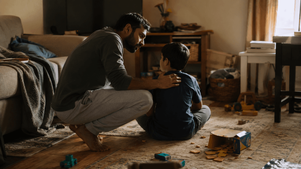 A South Asian dad sits face to face with his son on the living room floor, a few toys between them