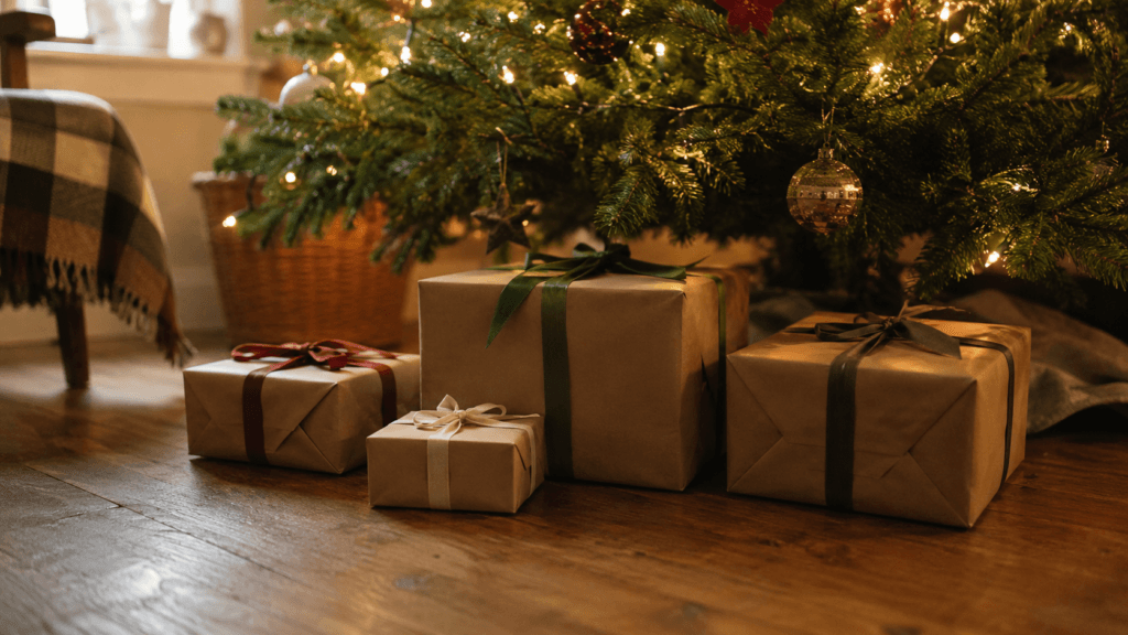Four wrapped presents of different sizes under a lit Christmas tree in soft morning light