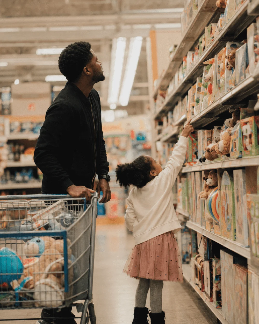 A parent and young child standing in a bright toy aisle looking up at a shelf of boxed toys