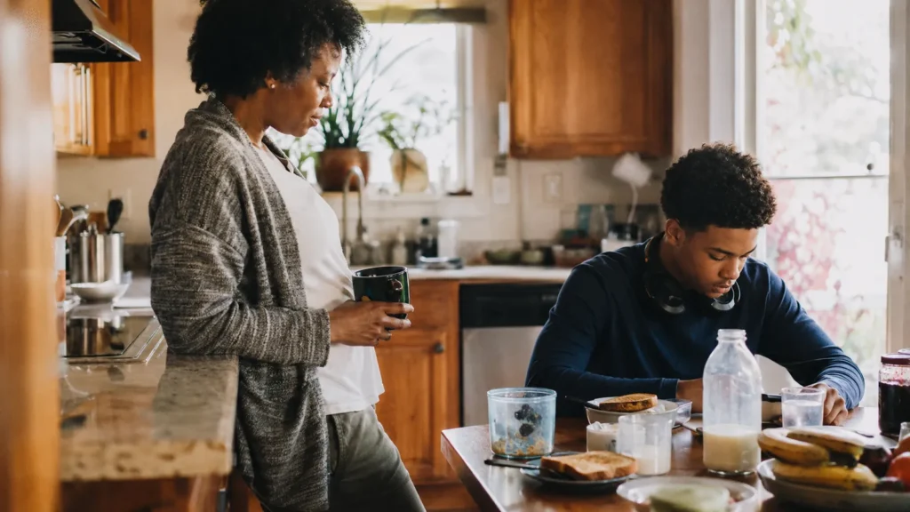 A tween sits at a kitchen island with headphones around their neck while a parent works nearby