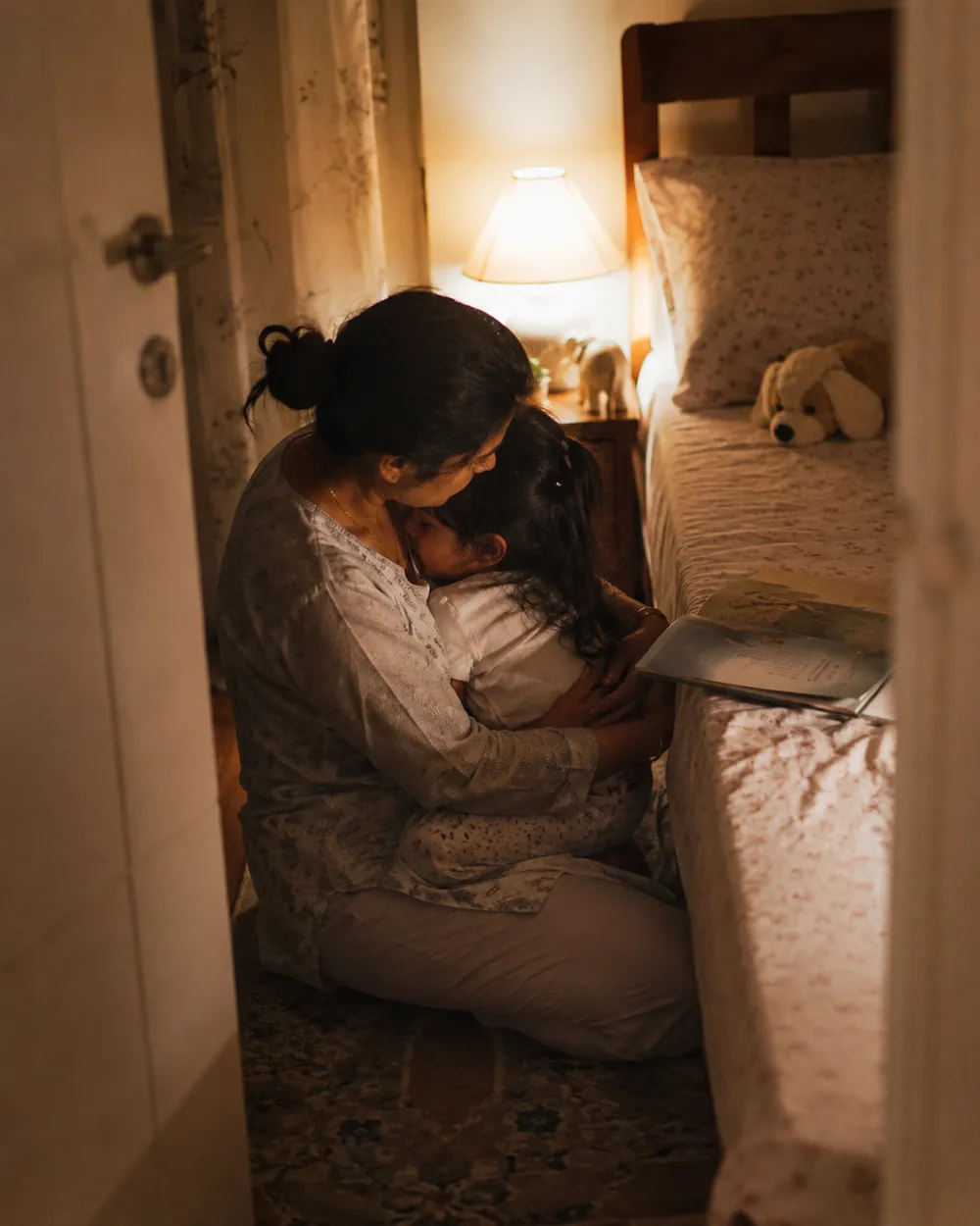 A mother hugs her daughter on the bedroom floor beside a lit night light at bedtime