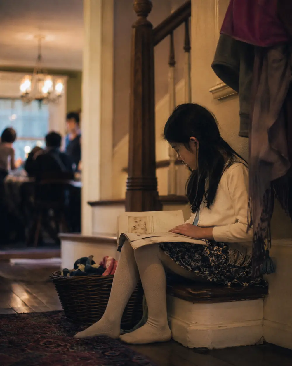 A child sitting alone on a staircase away from a noisy family gathering, holding a cup