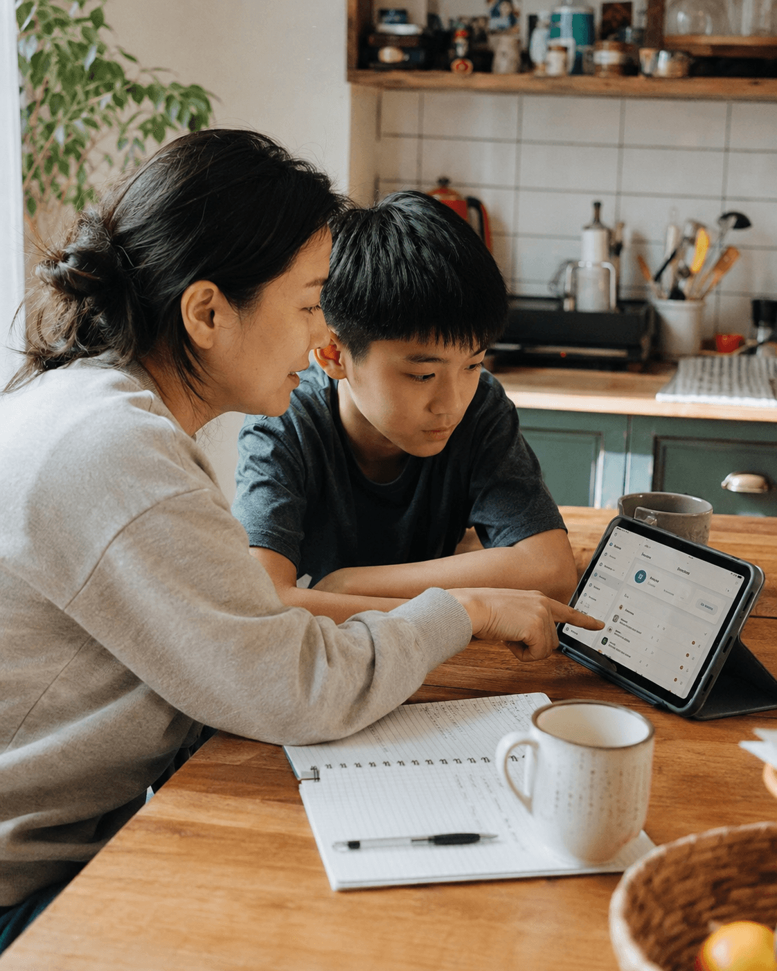 An Asian mother and her son at a kitchen table with a notebook, tablet and a mug between them