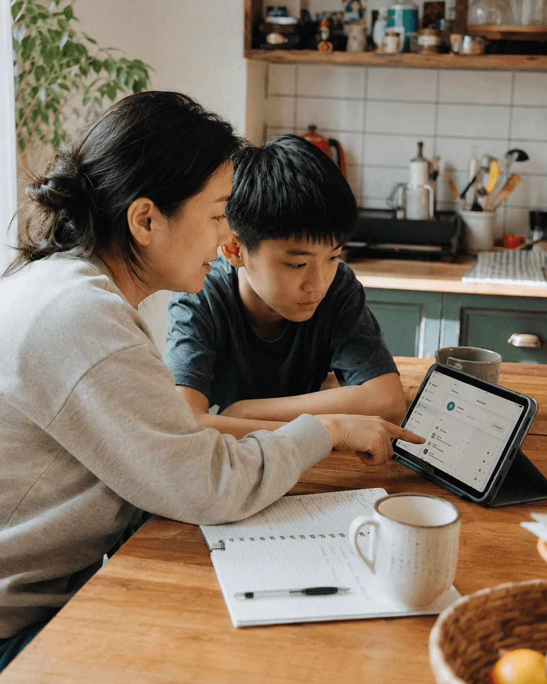 An Asian mother and her son at a kitchen table with a notebook, tablet and a mug between them