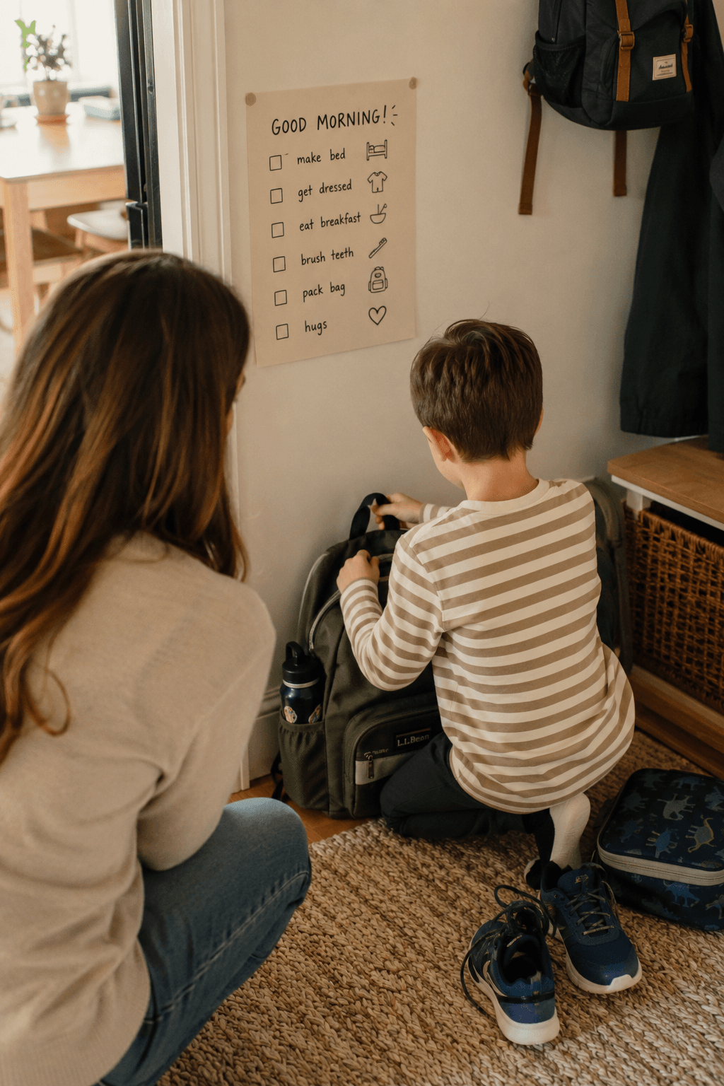 A parent crouching by the front door checking a morning checklist chart with their son