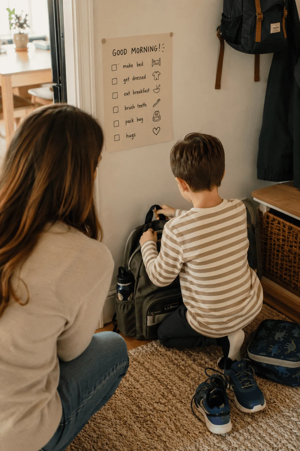 A parent crouching by the front door checking a morning checklist chart with their son