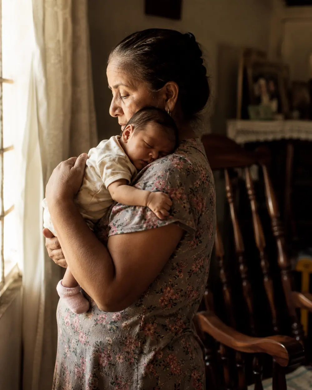 A woman standing at a bright window holding a sleeping newborn against her shoulder