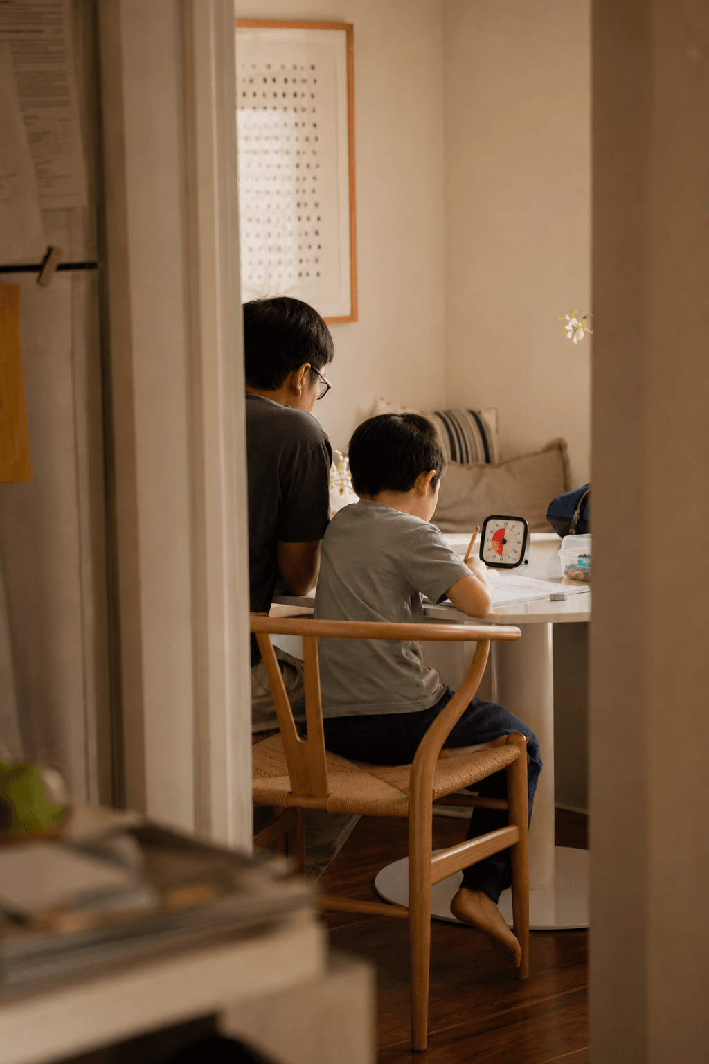 An Asian father sits with his son at the kitchen table doing homework with a visual timer beside them