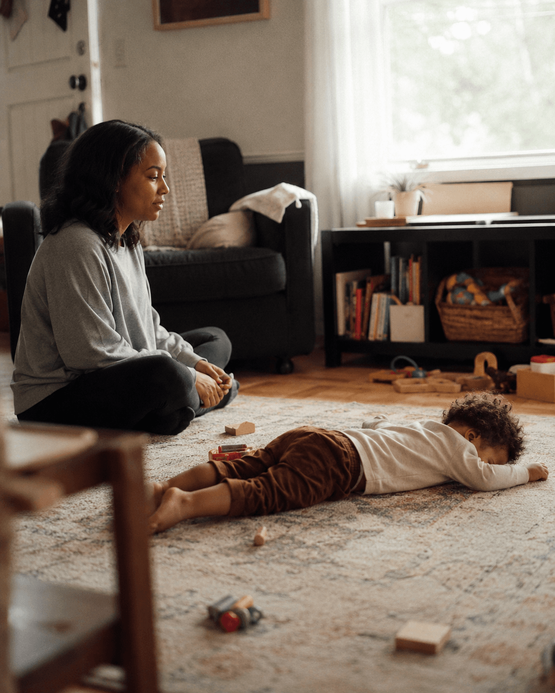 A Black mother sits calmly on the living room floor a short distance from her upset toddler and scattered blocks