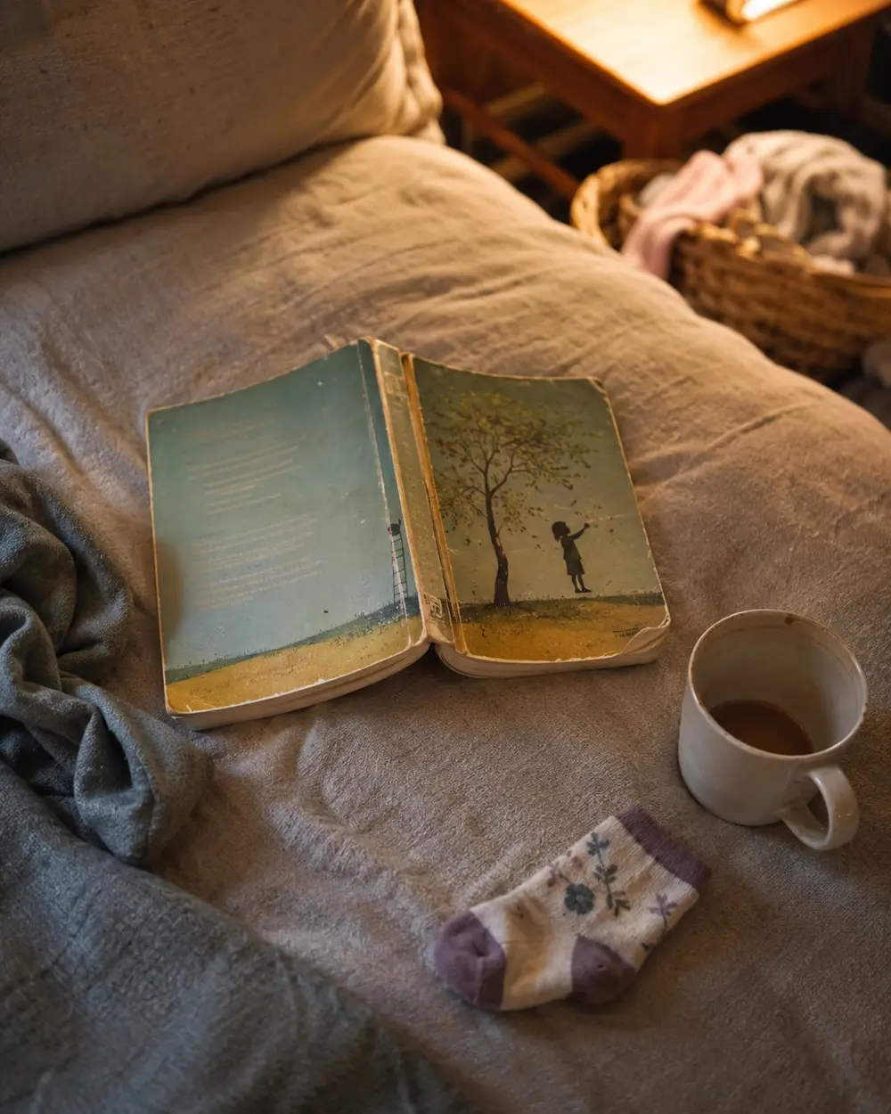 An unmade bed with an open picture book, a mug and a single baby sock resting on a soft throw