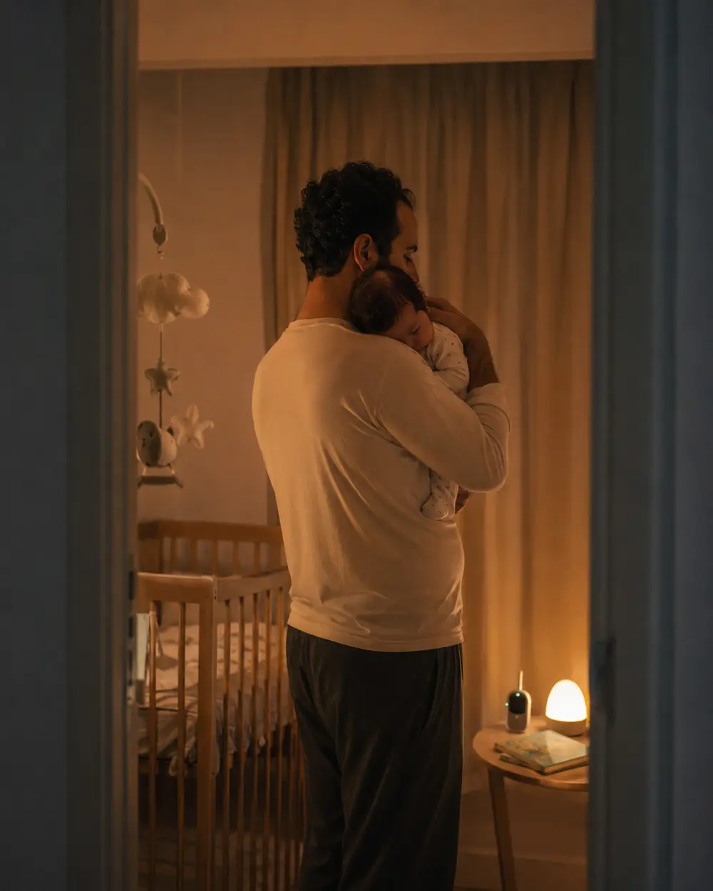A father standing beside a crib at night holding a newborn, a small night light glowing on the dresser