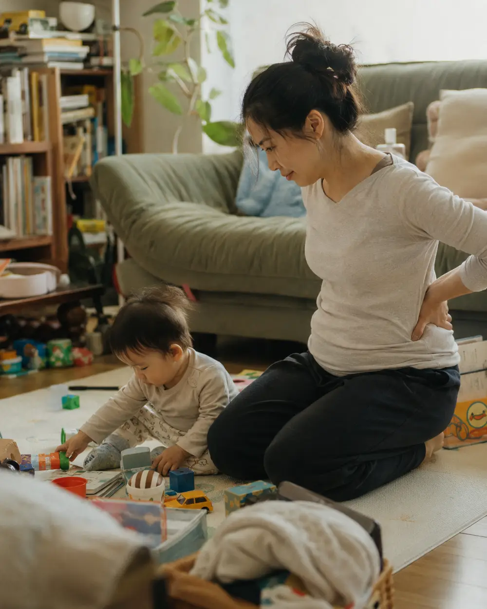 A mother sitting on the floor beside her baby, one hand pressed against her lower back