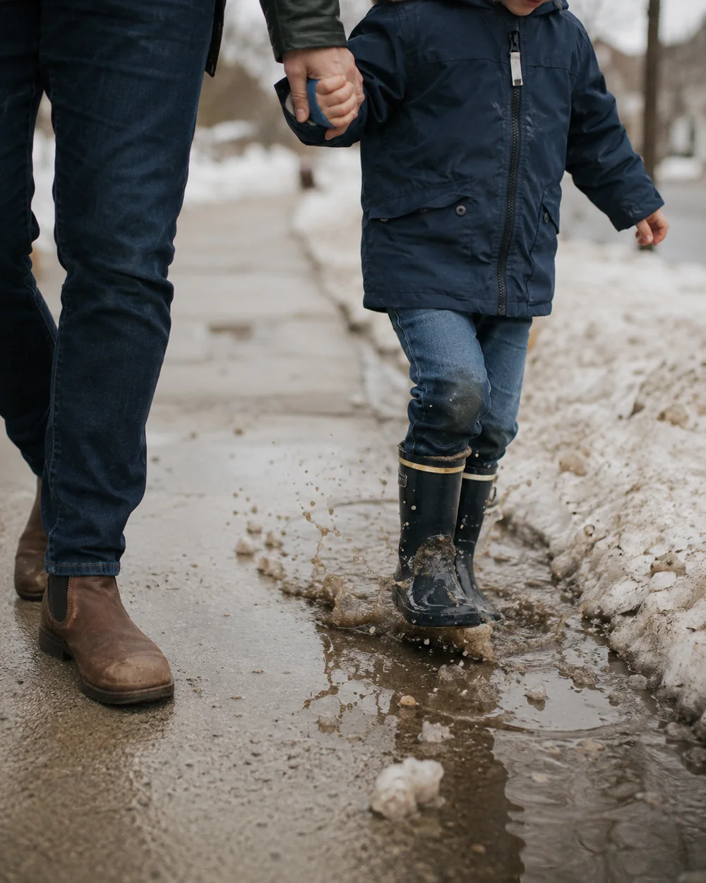 A child in boots walking home through slush on a grey street in late afternoon