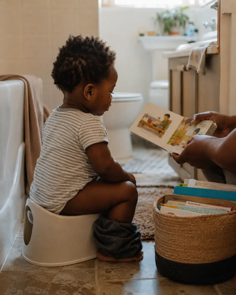 A toddler sits on a small potty in the bathroom with a short stack of board books within reach