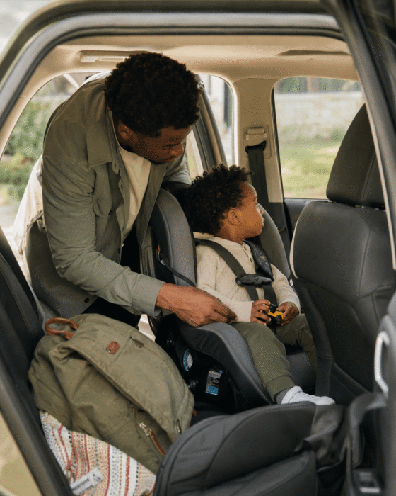 A Black father buckling his toddler into a car seat at morning dropoff, a small toy on the seat beside them