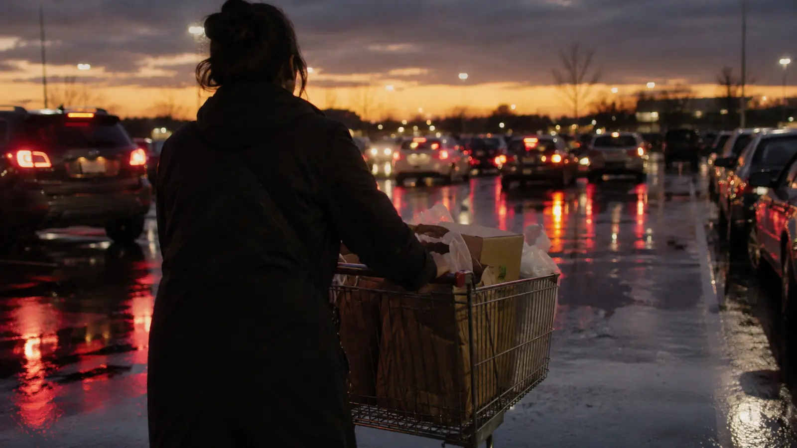A person pushing a cart across a wet parking lot at dusk with rows of cars and red tail lights