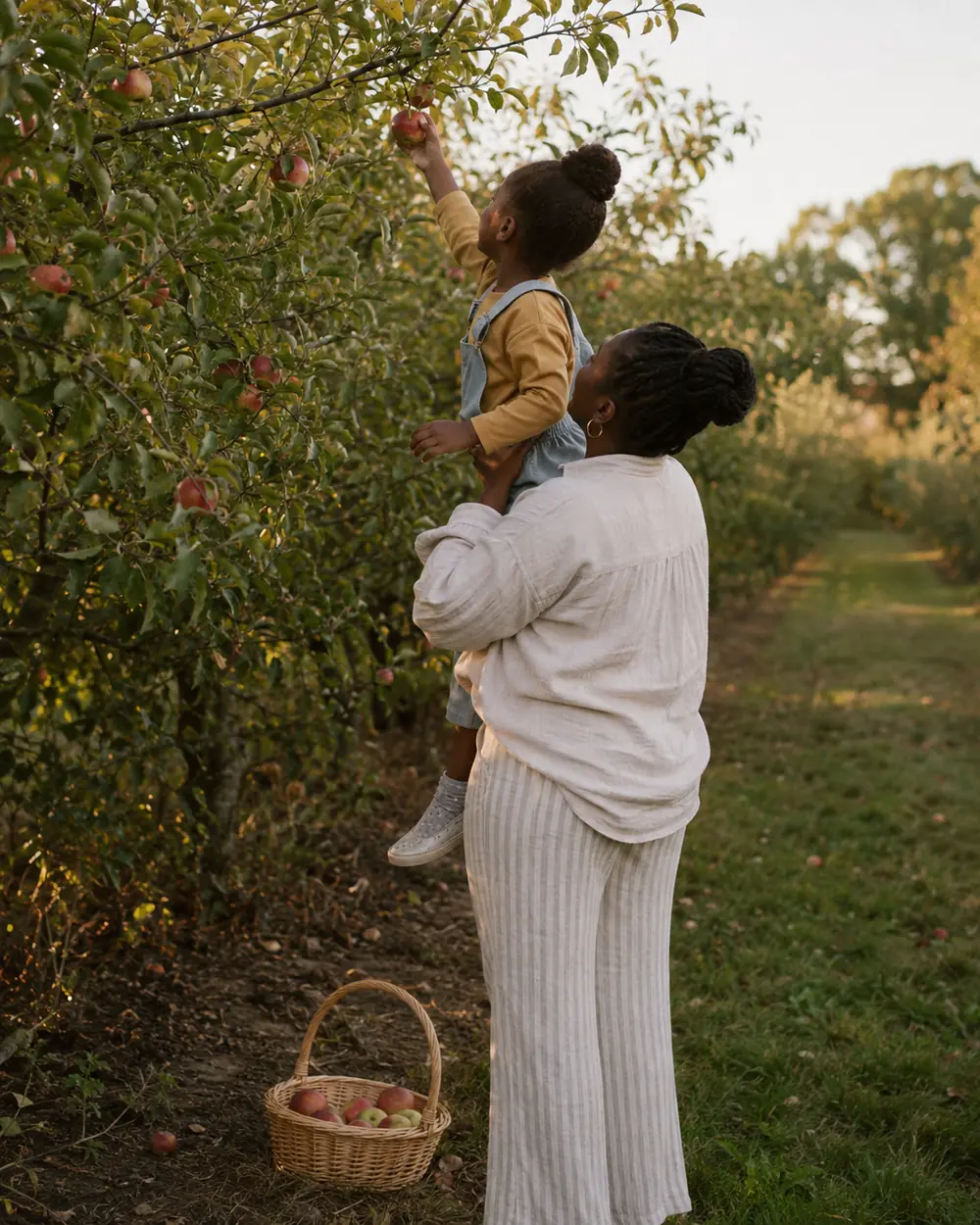 A mom lifts her young child to pick an apple from a tree in an orchard, with a basket on the grass