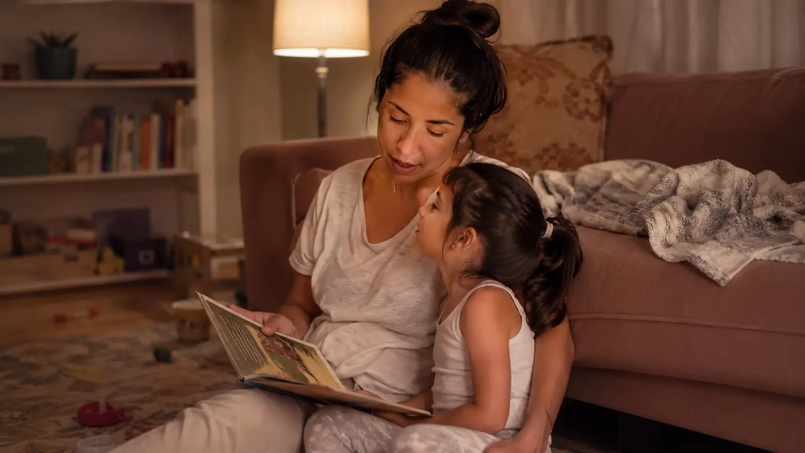 A parent and child reading an open picture book together on a lap in lamplight