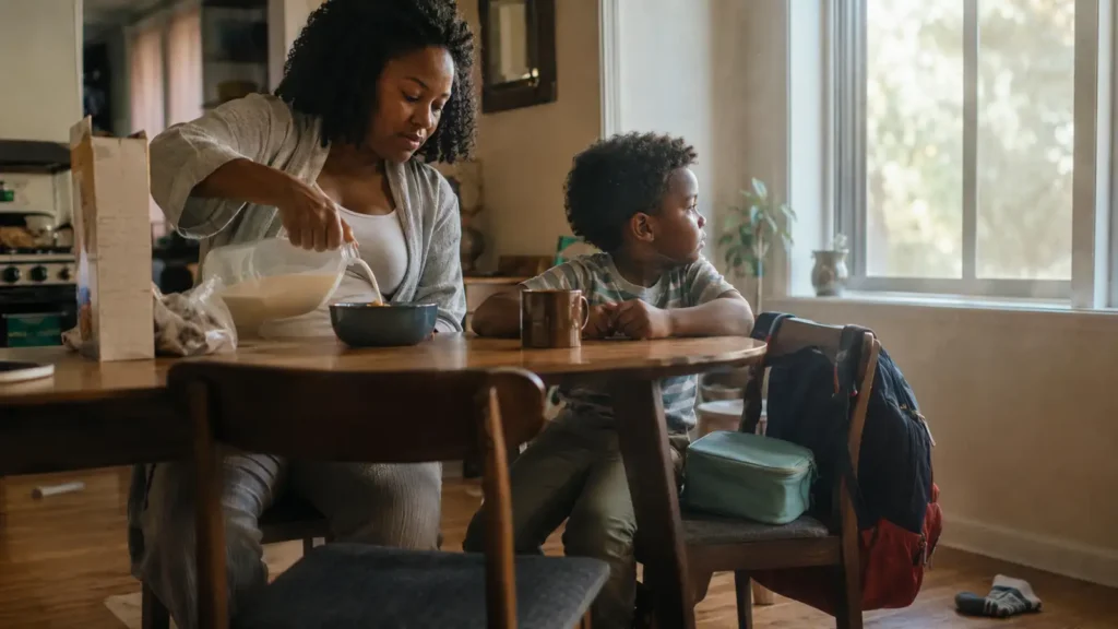 A mother and her son at a kitchen table in the morning, pouring milk, a school bag on the chair