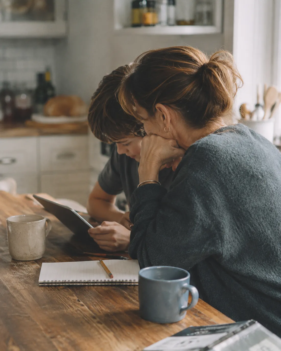 A mother and her son look at a tablet together at a wooden kitchen table, with a notebook, pencil and two mugs beside them