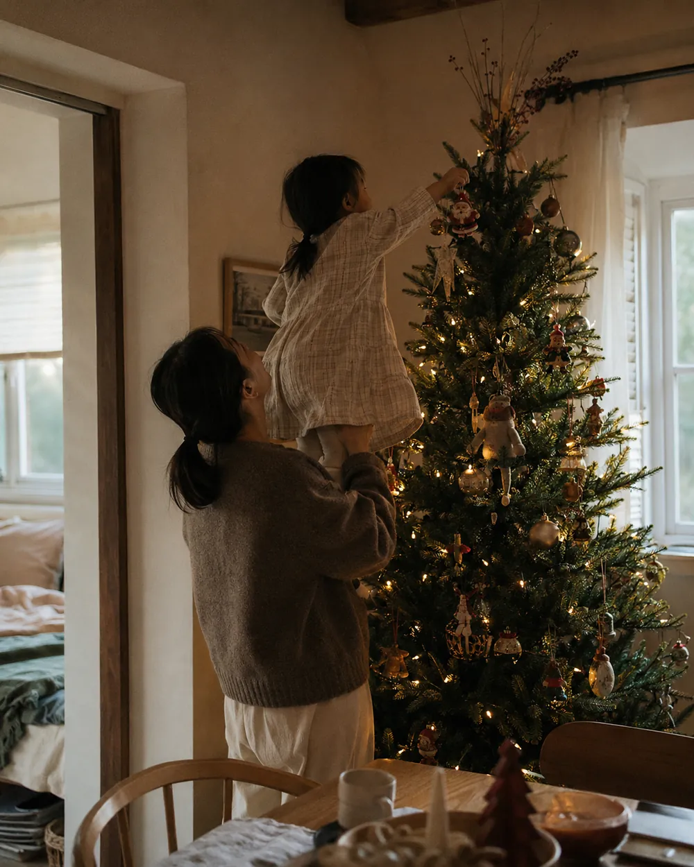 A mother lifts a toddler up to hang an ornament near the top of a Christmas tree