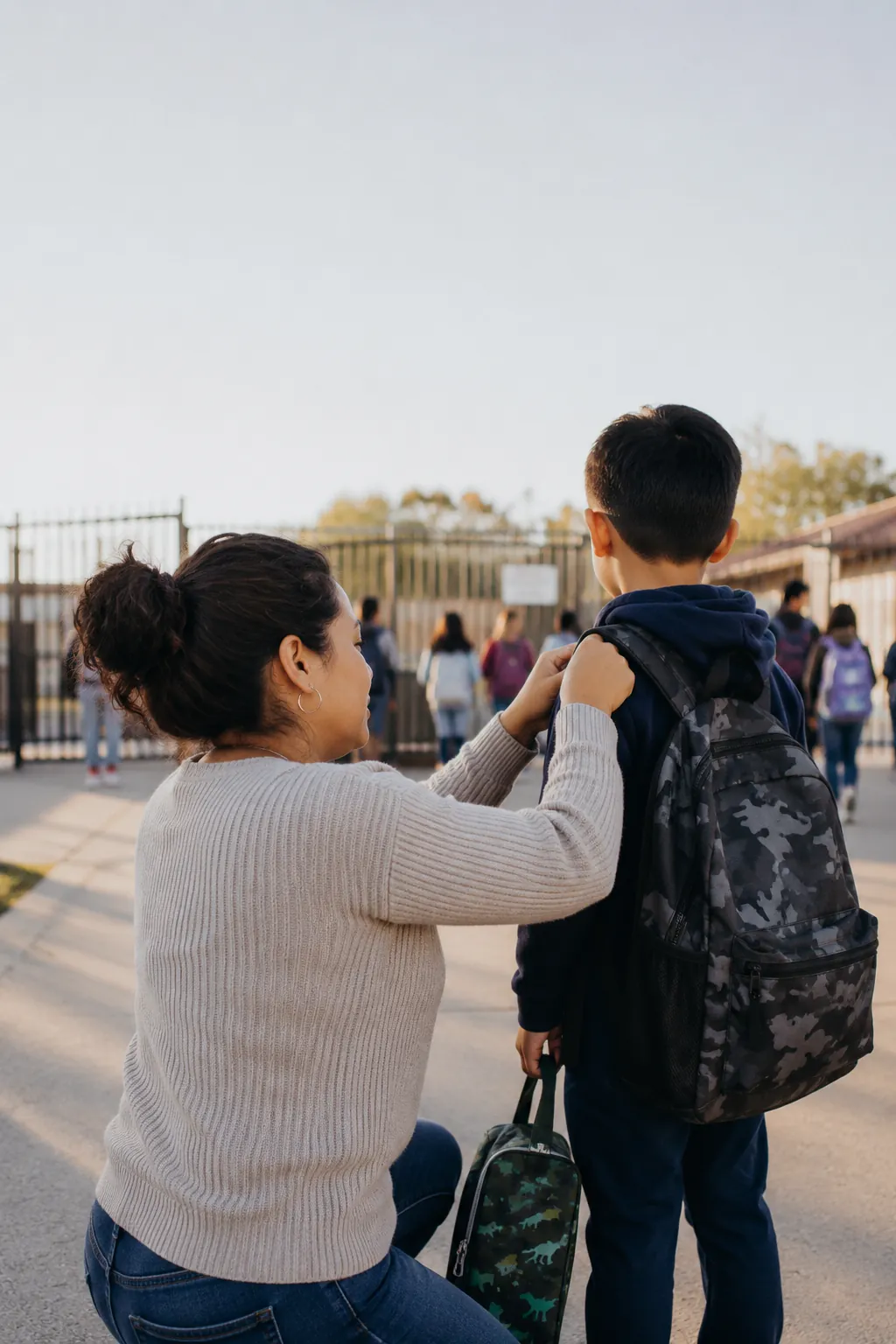 Mom adjusts her son backpack at the school gate on a normal drop-off morning