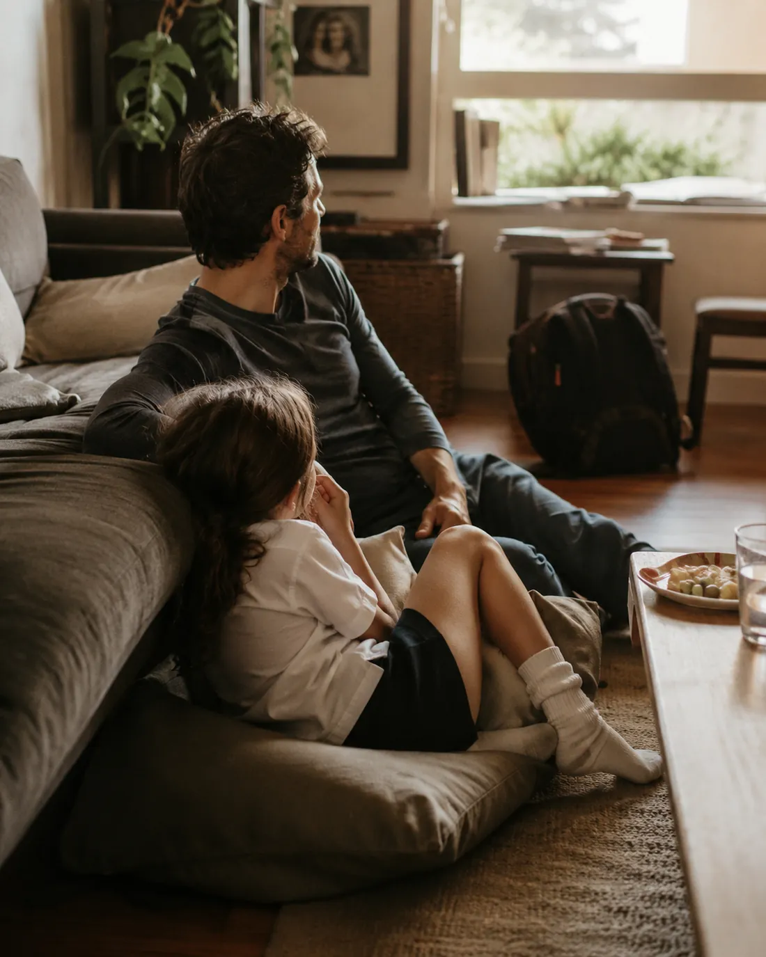 A dad and his daughter sit on the floor against the sofa after school, her backpack beside them